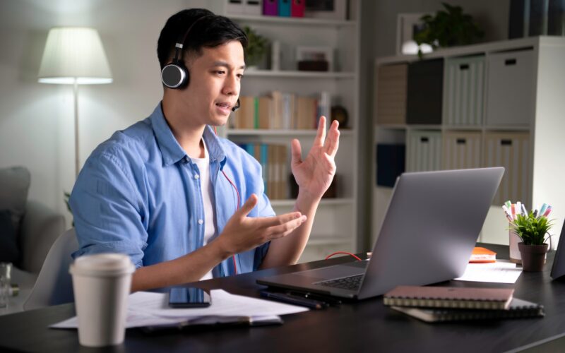 Focused young Asian business person, entrepreneur, speaker, student, office worker wearing headphones, looking at laptop screen and speaking in video conference call, giving lecture, joining online class using a laptop when work from home.
